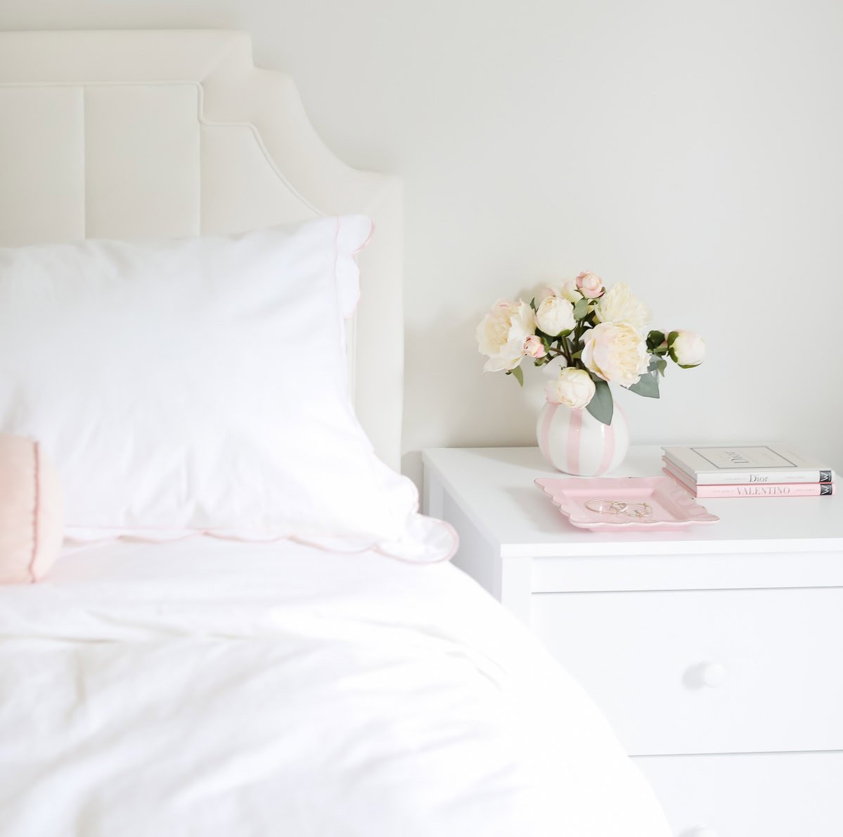 A white bed with a white pillow and blanket next to a white nightstand holding a vase of white flowers, a pink tray, and a small stack of books creates the perfect Pottery Barn look for less.