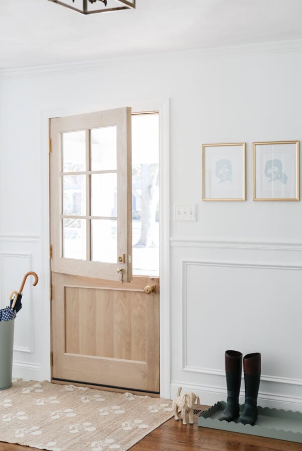 A wooden door with glass panels is partially open in a white entryway, showcasing a Pottery Barn look for less with an umbrella stand, doormat, rubber boots, and two framed pictures.