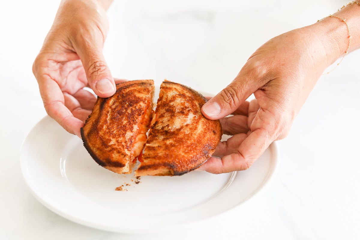 Hands holding a sliced, toasted campfire pizza sandwich over a white plate.