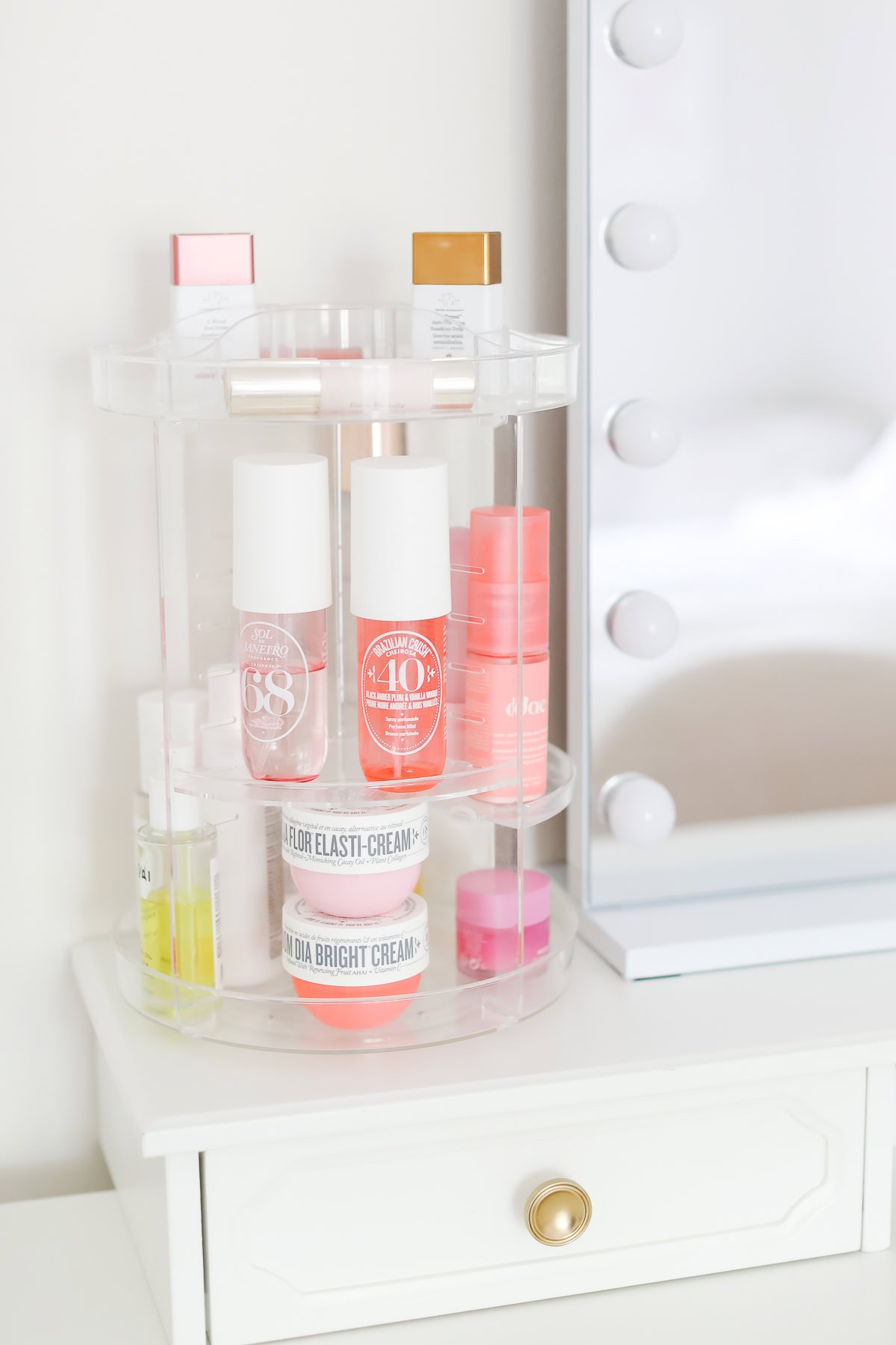 A clear acrylic rotating organizer holds various skincare and beauty products on a white vanity next to a mirror with lights, making it the perfect addition to refresh your space this Prime Day.