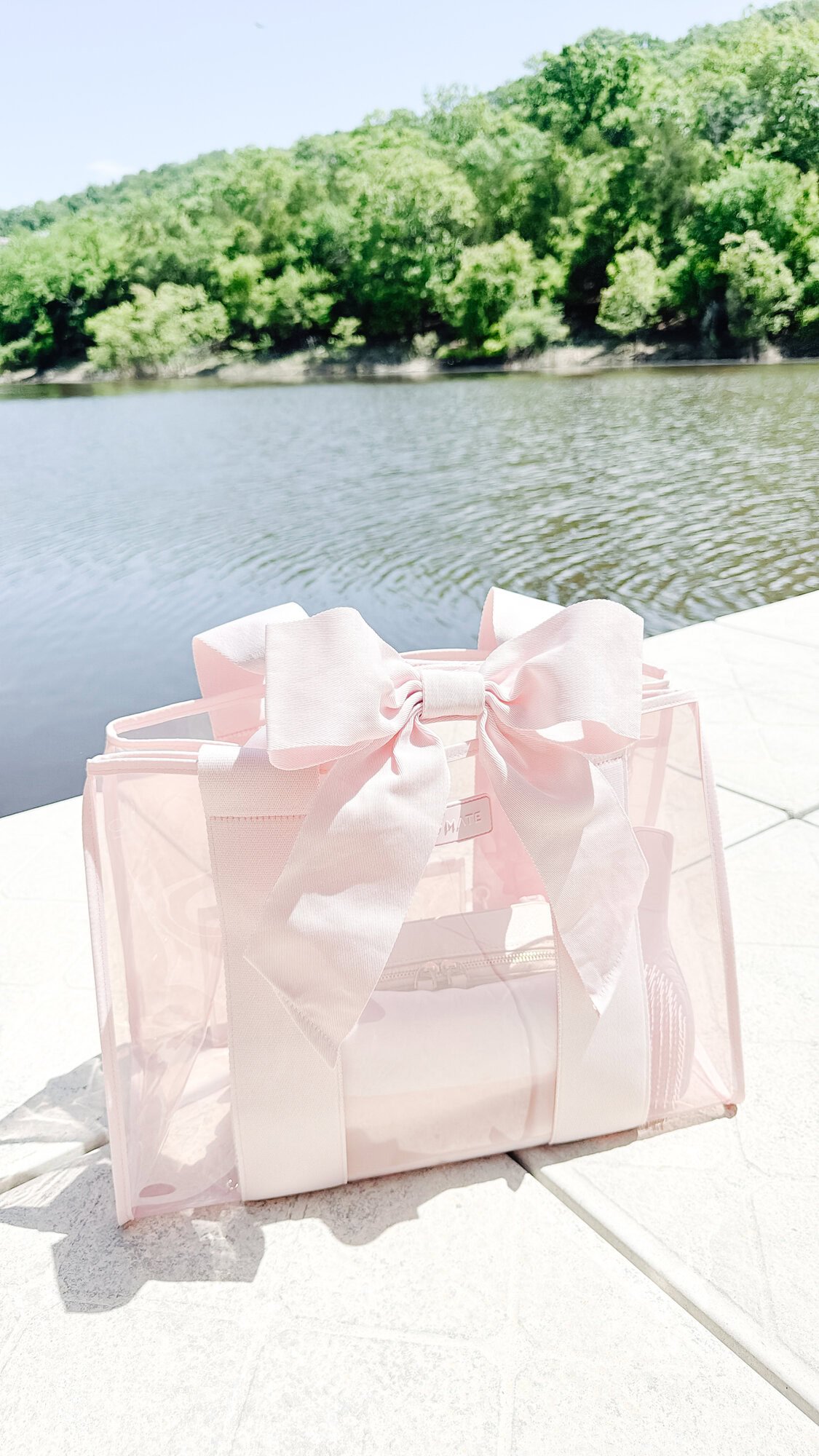 A transparent pink bag with large bows sits on a white patio next to a body of water and green trees, perfect for showing off your style this Prime Day.