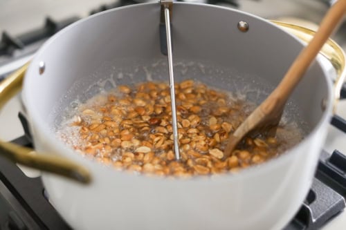 Peanut brittle preparation in progress: nuts boiling in a pot on the stovetop, accompanied by a thermometer and wooden spoon for precise cooking.