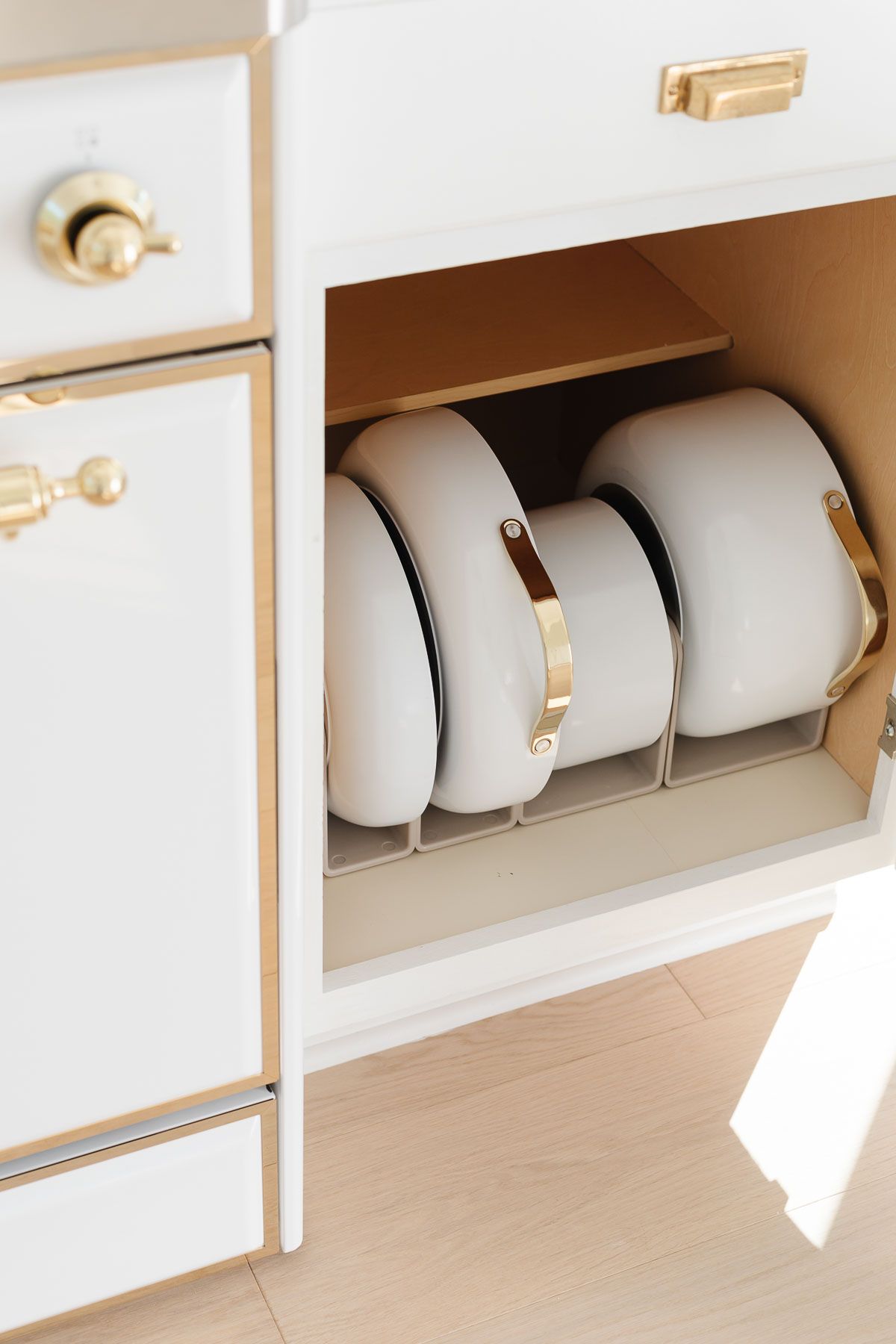 White pots with gold handles are neatly stored upright inside a lower kitchen cabinet with a built-in rack—perfect for organizing your space before Prime Day. The cabinet and appliances are white with gold accents.