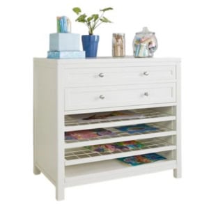 A white chest with two drawers and four open shelves, perfect for craft space ideas, holds art supplies on top alongside jars and potted plants.