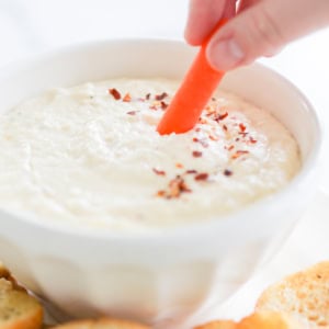 Hand dipping a carrot stick into a bowl of creamy Asiago cheese dip topped with chili flakes, surrounded by toasted bread slices.