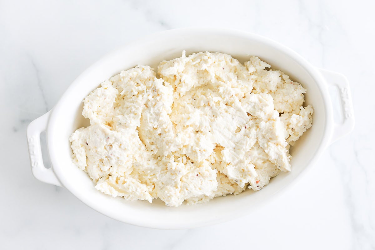 A white oval dish filled with a creamy, textured Asiago Cheese Dip sits on a marble surface.