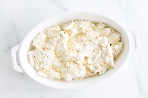 A white oval dish filled with Asiago Cheese Dip, revealing a creamy, textured mixture resting on a marble surface.