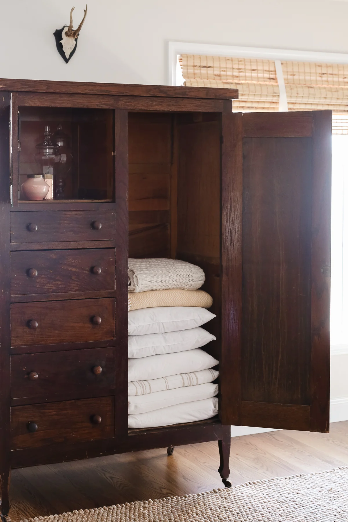 An open wooden cabinet with drawers and shelves, perfect for organizing crafts, contains folded blankets and a glass bottle. Above it, a small mounted animal head adds a touch of rustic charm.