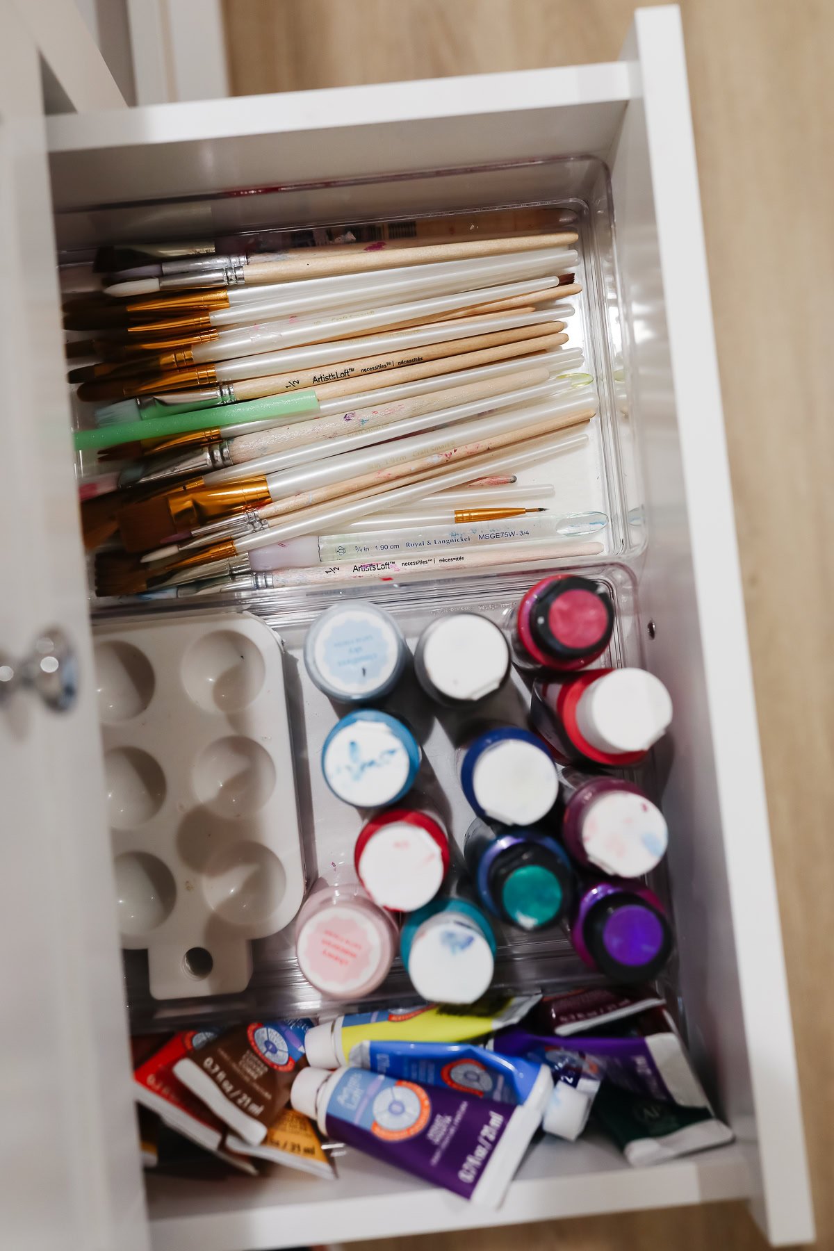 An open drawer cleverly organizing crafts features paintbrushes, various tubes and bottles of paint, and a perfectly placed paint palette.