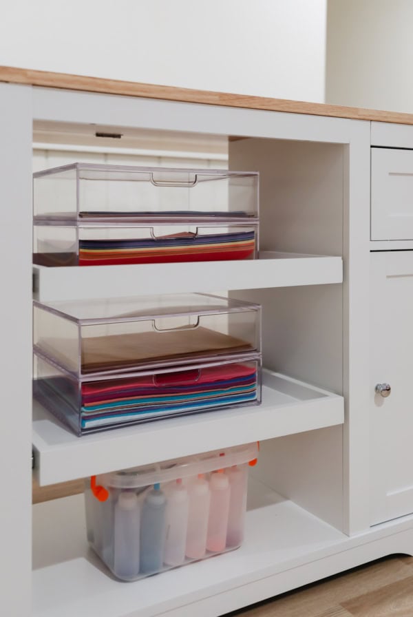 White cabinet with open shelves displaying transparent boxes filled with colorful papers and organizing crafts supplies, alongside storage bins containing spray bottles.