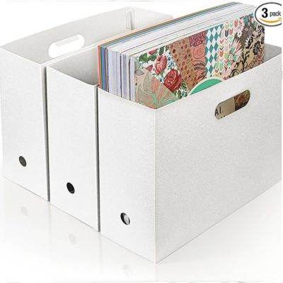 Three white storage boxes designed for organizing crafts, one filled with colorful patterned papers, are displayed on a reflective surface.