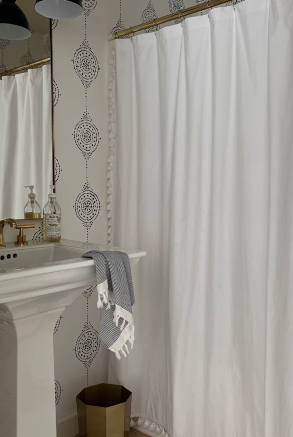 The bathroom features a white shower curtain, gold fixtures, and a pedestal sink. A gray towel hangs on the sink, next to a gold trash bin on the floor. Serena and Lily wallpaper with a patterned design adds a touch of elegance to the space.