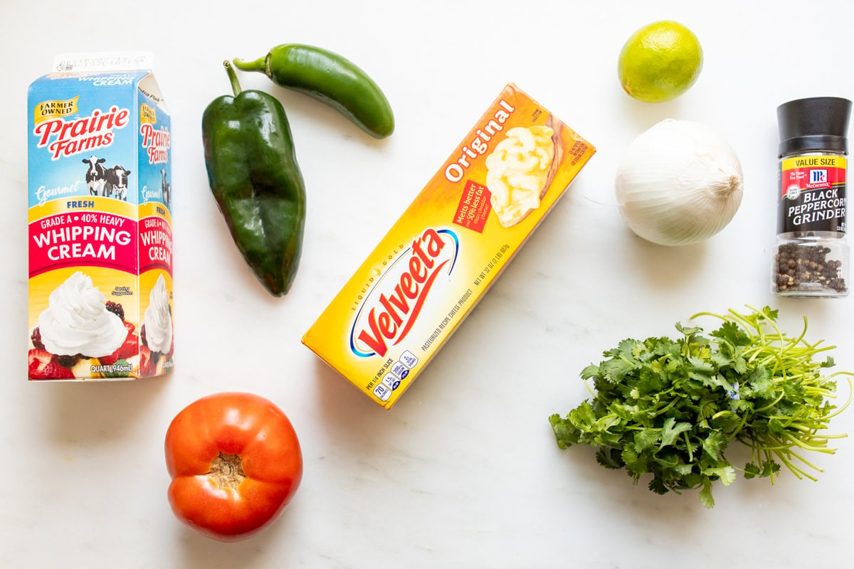 Ingredients on a countertop include whipping cream, Velveeta cheese ready for a creamy dip, a tomato, poblano pepper, jalapeño, lime, onion, black pepper grinder, and cilantro.