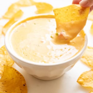 A hand dips a tortilla chip into a bowl of creamy Velveeta cheese dip, surrounded by crispy tortilla chips.