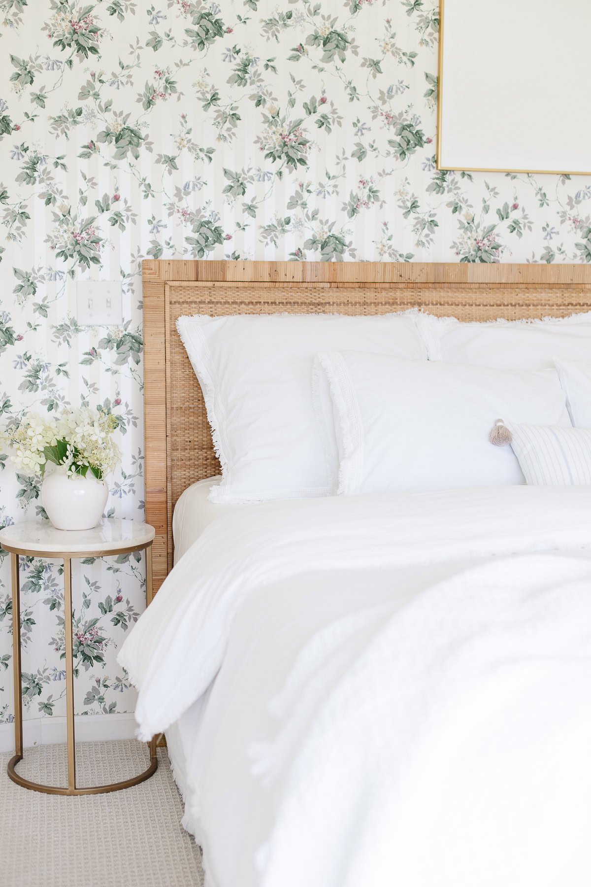 A neatly made bed with white bedding and a wicker headboard offers a Serena and Lily look for less, paired with a round side table holding a white vase of flowers, set against floral wallpaper.