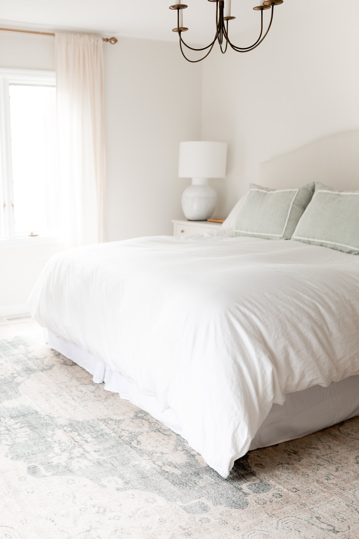 A bright bedroom with a neatly made bed, light-colored linens, a bedside table with a lamp, a window with curtains, and a patterned rug on the floor—perfect for showcasing your ideal rug size or consulting a rug size guide.