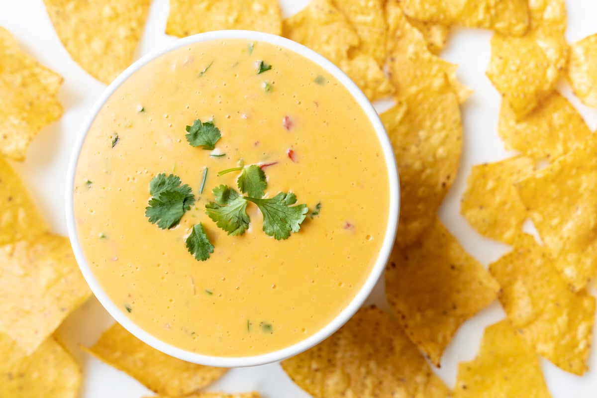 A bowl of velveeta cheese dip with a cilantro garnish, surrounded by crunchy tortilla chips on a pristine white surface.