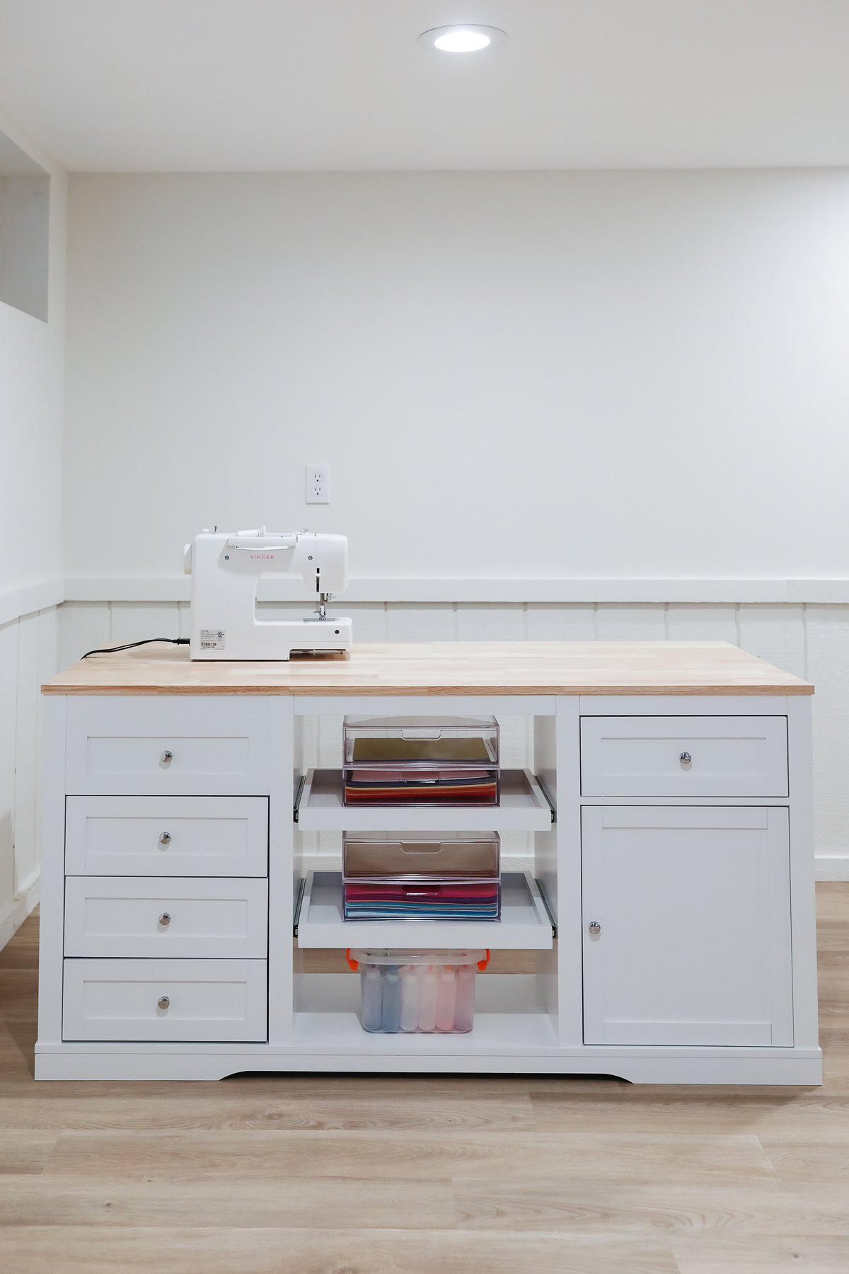 A white sewing table with a wooden top anchors this craft space, housing a sewing machine, drawers, and shelves filled with colorful vinyl. Storage boxes neatly fit underneath, while the room's white walls and wood flooring complete the organized craft space ideas.