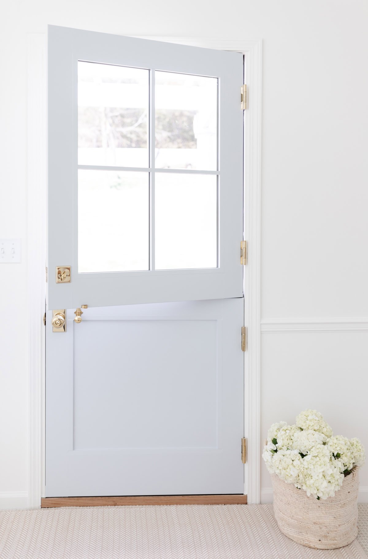 A light blue Dutch door with brass hardware is half open, letting in sunlight. A basket of white flowers sits on a cozy rug—perfectly chosen with a handy rug size guide—to welcome guests warmly.