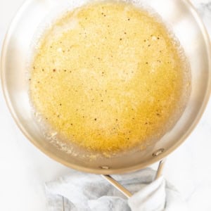 Top view of a steel frying pan containing melted browned butter with small bits, hinting at the rich aroma of garlic parmesan sauce, and a white towel on the handle.