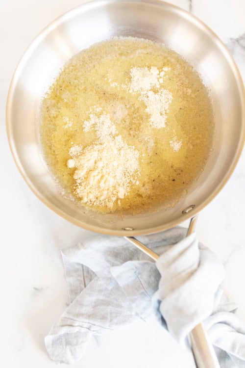 Flour and melted butter in a stainless steel pan on a marble surface, with a light cloth wrapped around the handle, ready to create a rich garlic parmesan sauce.