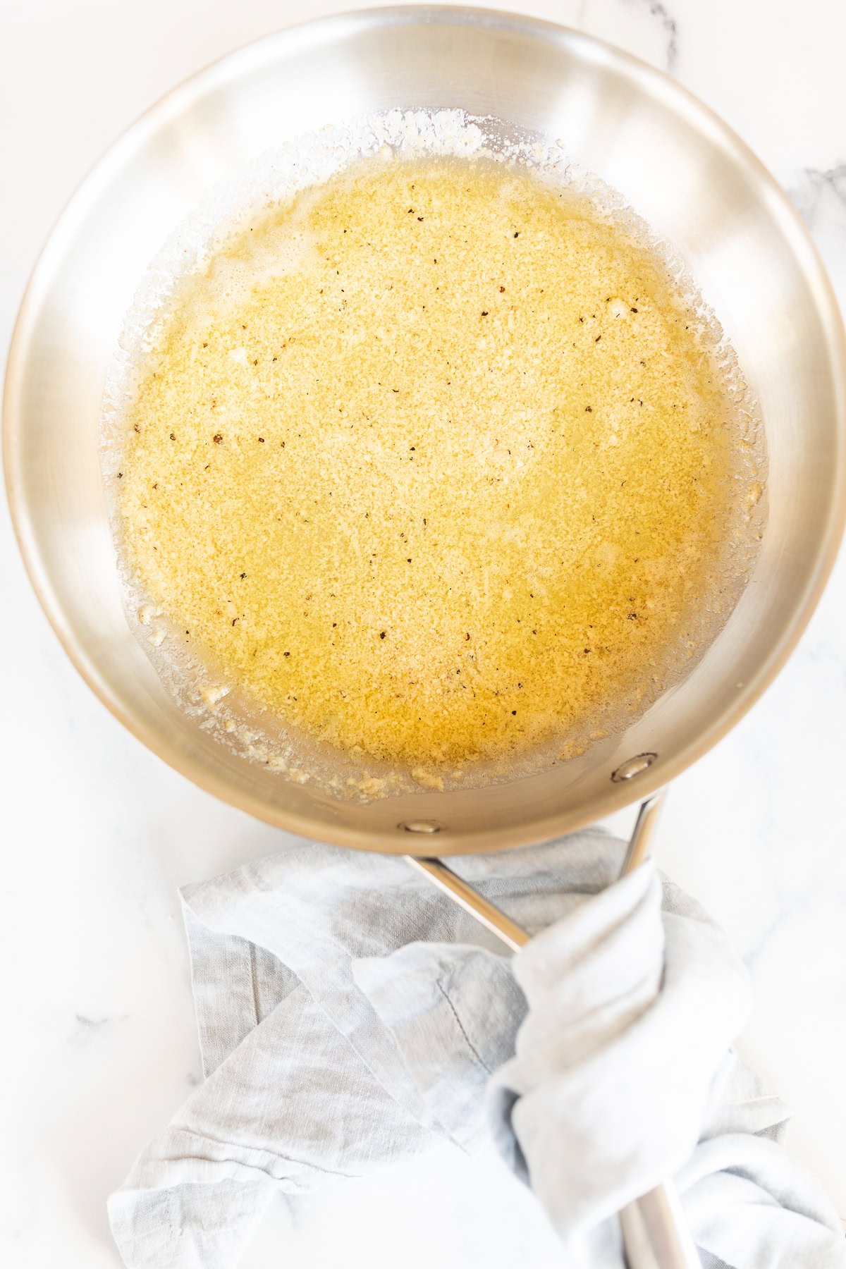 A stainless steel pan with melted butter and a hint of garlic parmesan sauce, showing a light golden-brown color, rests on a marble countertop with a cloth wrapped around the handle.