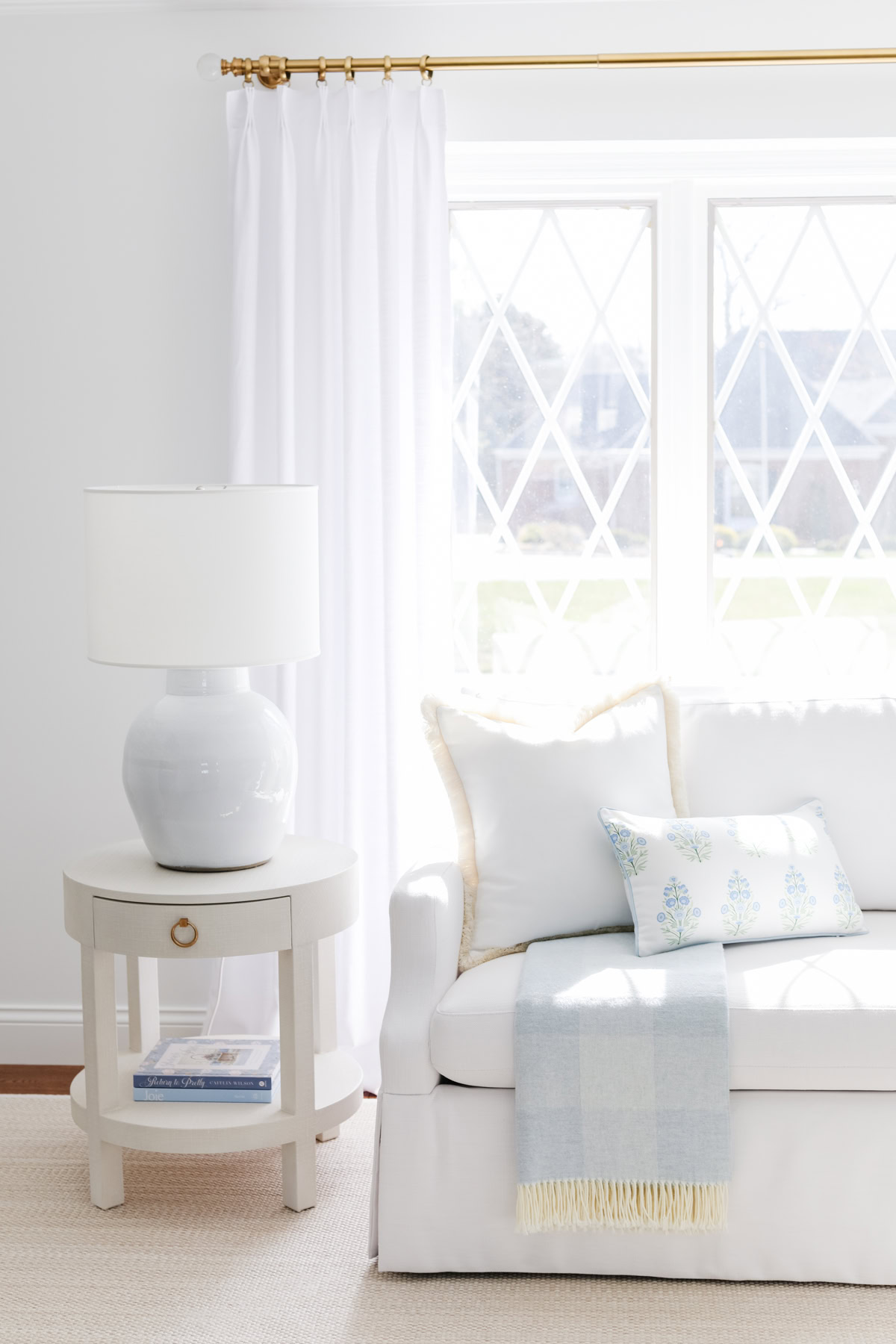 A round side table with a white lamp sits next to a white sofa with light blue pillows and a throw, in front of a large window with white curtains—an inviting setting to consult your rug size guide for the perfect finishing touch.