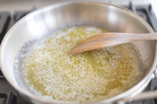 Melted butter in a stainless steel pan is being stirred with a wooden spoon, creating the base for a rich garlic Parmesan sauce.