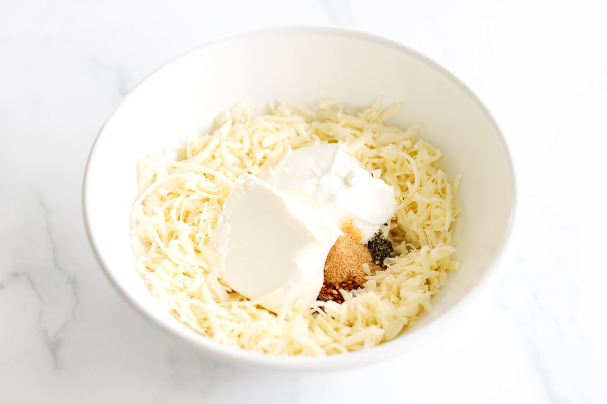 A bowl featuring Asiago Cheese Dip alongside shredded cheese, sour cream, and various spices rests on a pristine white surface.