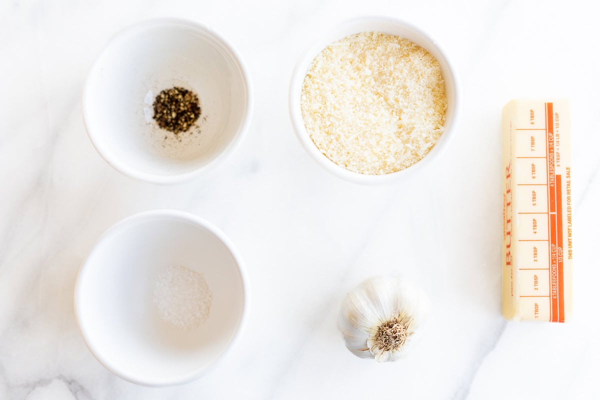 Salt, pepper, and grated cheese sit in bowls on a white marble surface alongside a bulb of garlic and a stick of butter—the essential ingredients for crafting a savory garlic parmesan sauce.