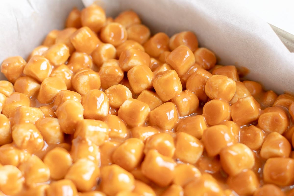 A tray filled with small, shiny caramel-coated cubes rests on parchment paper, featuring delightful Peanut Butter Marshmallow Squares.