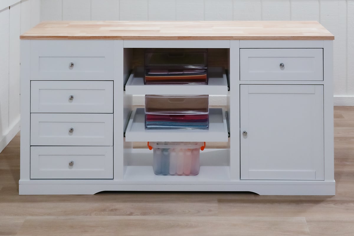 A white storage cabinet with a wood top offers clever craft space ideas. It features multiple drawers and shelves holding paper and transparent storage bins, perfectly organized. Positioned on a wooden floor in a room with white paneled walls, it blends functionality with style.