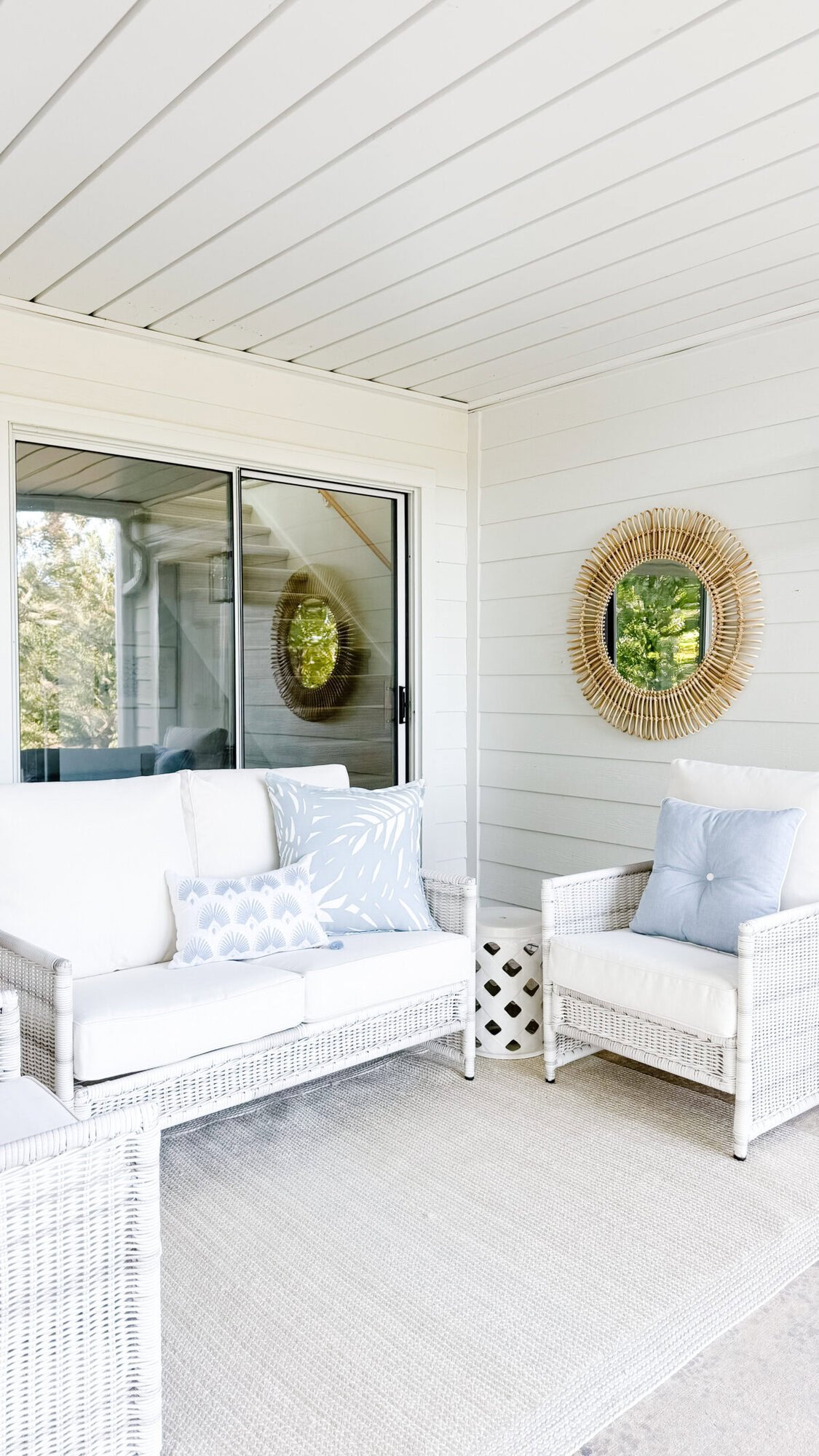 A white wicker outdoor seating area with a sofa, armchair, light blue and white pillows, a neutral rug, and a round rattan mirror on a white-paneled wall offers a chic Serena and Lily look for less.
