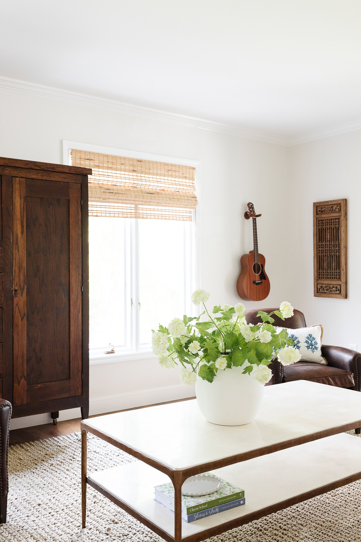 A bright living room with a white coffee table, a vase of white flowers, a wooden cabinet, a guitar on the wall, and a window with a bamboo shade—perfect for showcasing your rug size or consulting our rug size guide.