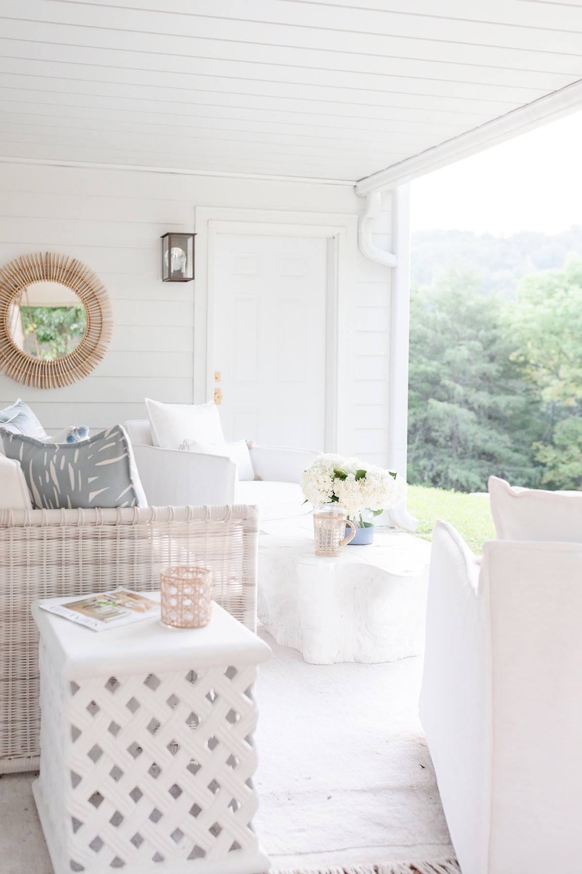 A bright, white outdoor patio with wicker furniture, white cushions, a small table with flowers, and a mirror on the wall offers a serena and lily look for less, complemented by a lovely view of greenery outside.