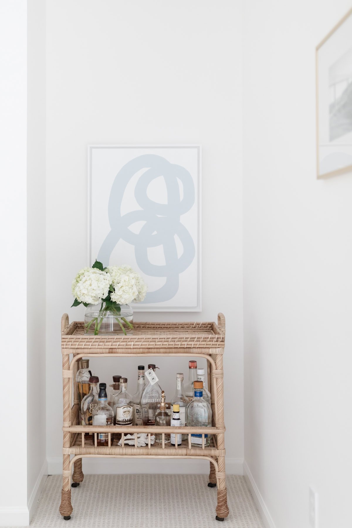 A wicker bar cart with assorted liquor bottles, a bouquet of white hydrangeas in a vase, and abstract art on the wall brings a bright, minimalist room together—achieve this Serena and Lily look for less.