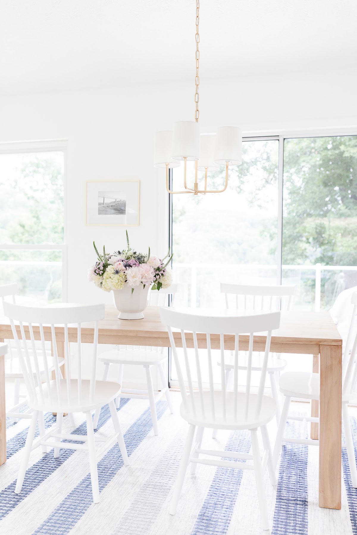 Bright dining room with white chairs and a wooden table topped with a flower arrangement, under a modern chandelier, large windows, and a blue-striped rug for that Serena and Lily look for less.