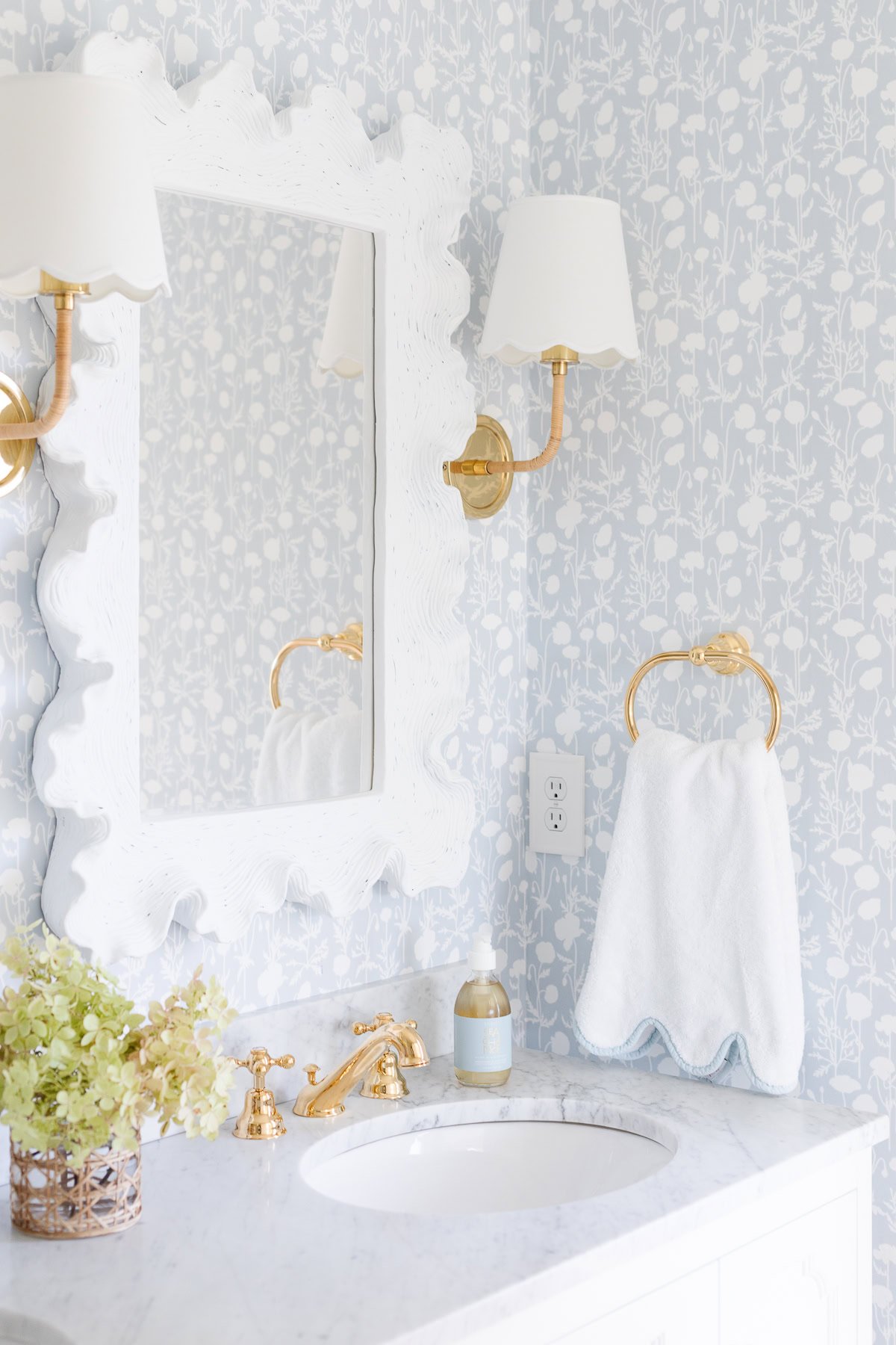 A bathroom with a white marble sink, gold faucet, and Serena and Lily floral wallpaper. A white framed mirror and dual wall lamps are above. A gold towel ring holds a white towel next to the mirror.