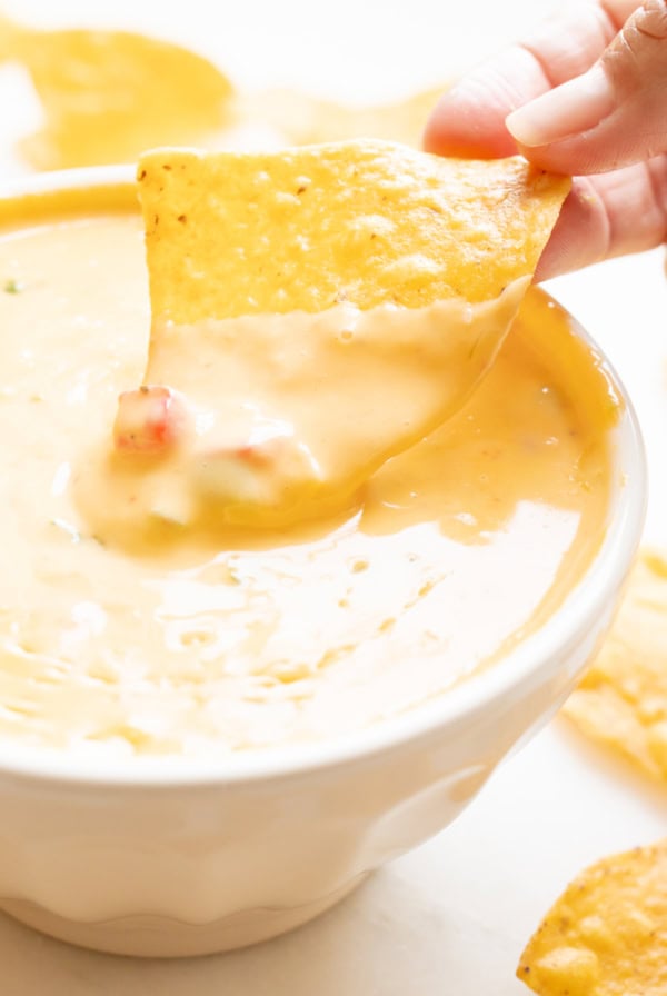 A hand dips a tortilla chip into a bowl of velveeta cheese dip, enriched with small vegetable pieces.