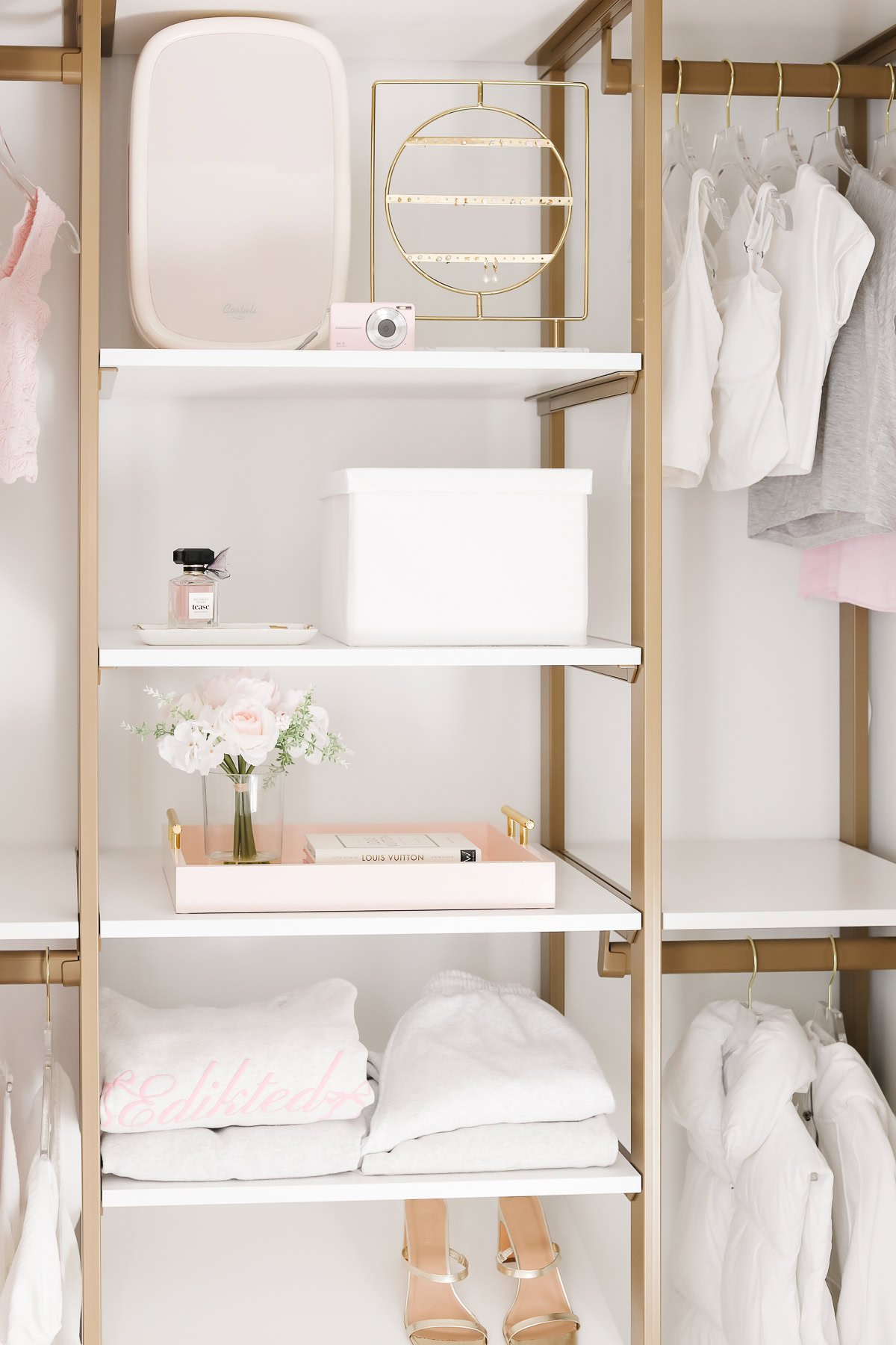 Organized kids closet with gold shelving, featuring pink flowers, a white storage box, perfume, clothes hangers, and folded white garments and towels.