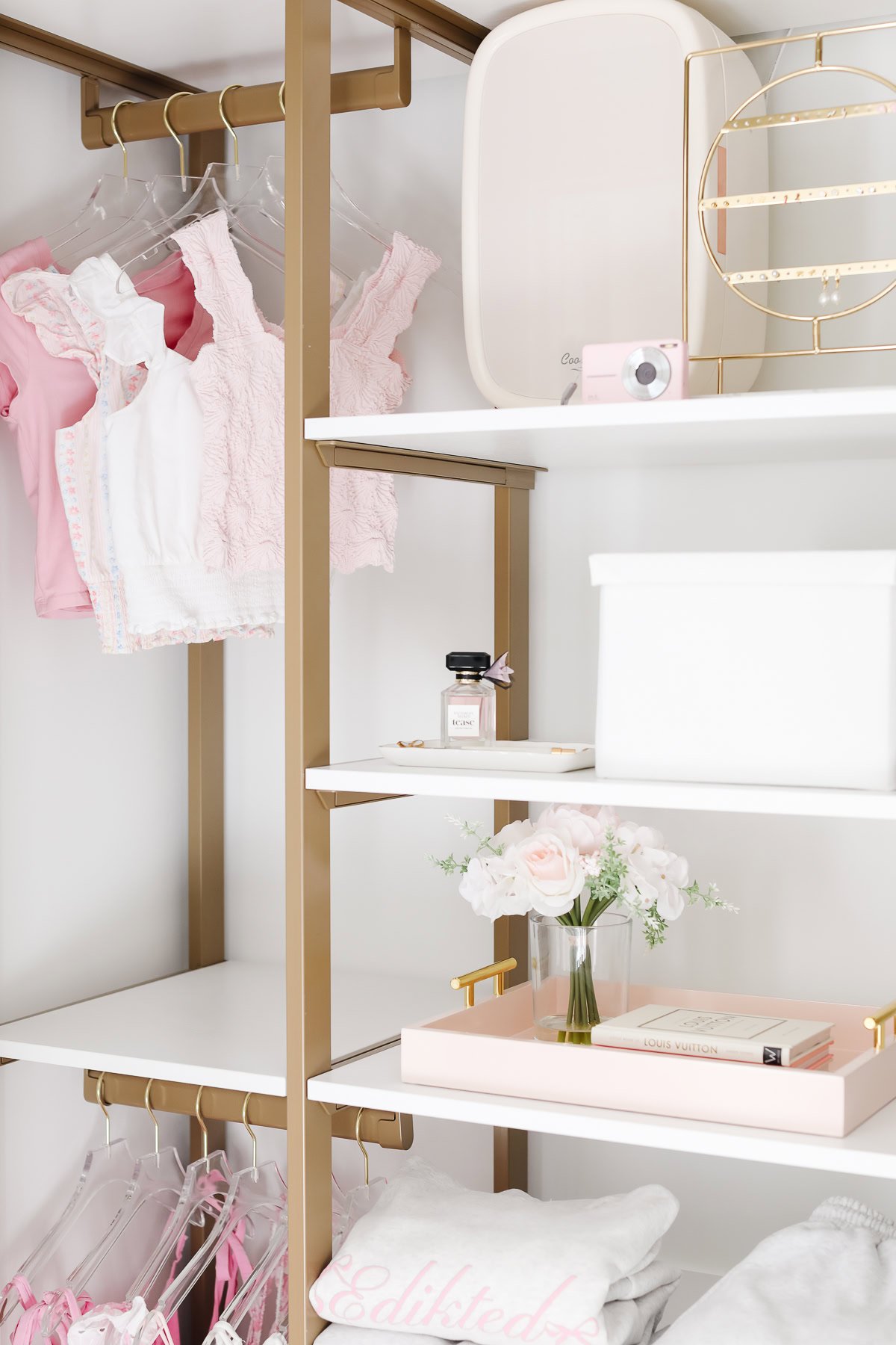 In a meticulously organized kids closet, gold shelves display hanging pink and white tops. A clear storage box holds essentials, while a small perfume bottle sits alongside white flowers in a vase and charming decorative items.