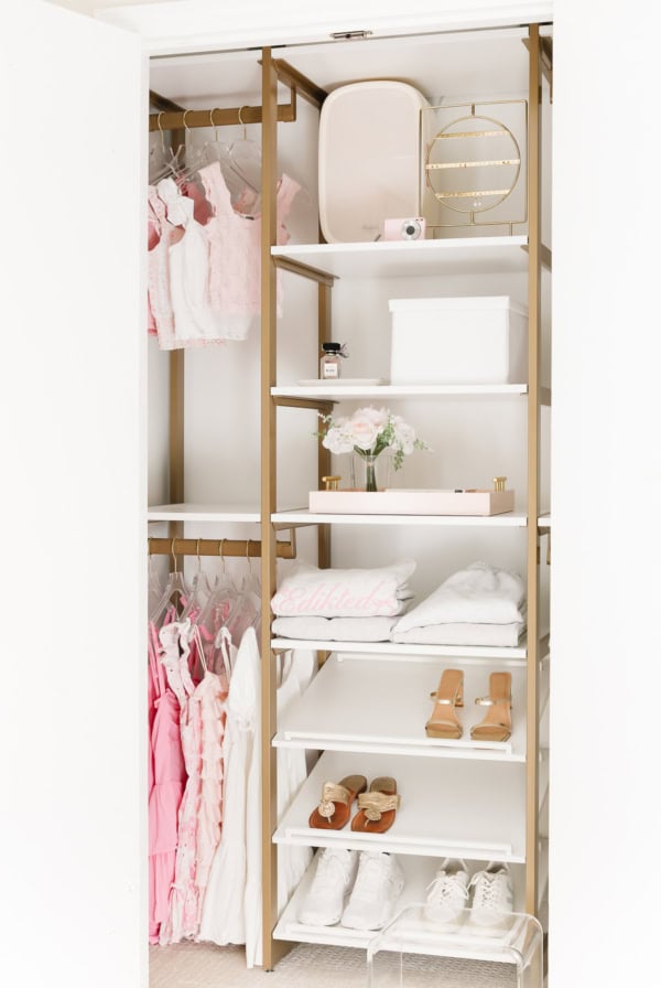 An open kids closet with organized shelves holding shoes, bags, a camera, and playful decor. To the left, clothes hang neatly featuring pink, white, and patterned garments that add a splash of fun.