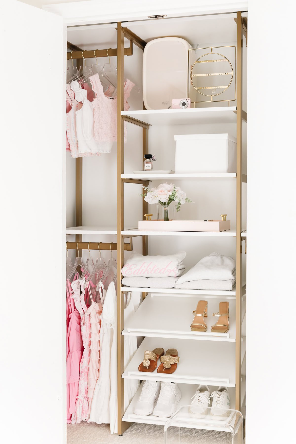 An open kids closet with organized shelves holding shoes, bags, a camera, and playful decor. To the left, clothes hang neatly featuring pink, white, and patterned garments that add a splash of fun.