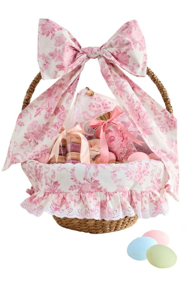 A wicker basket decorated with pink floral fabric and a large bow, filled with pastel-colored treats and Easter basket fillers, with three pastel candies placed beside it.