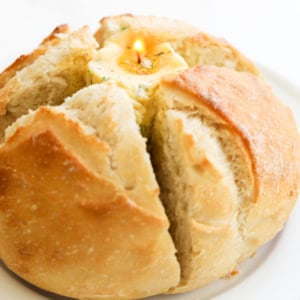 A round loaf of bread with the top partially sliced open, revealing a lit candle placed inside a dollop of spread on a plate.