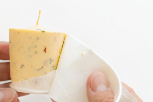 A person peeling a white paper wrapper off a wedge of seasoned cheese with visible herbs and spices.