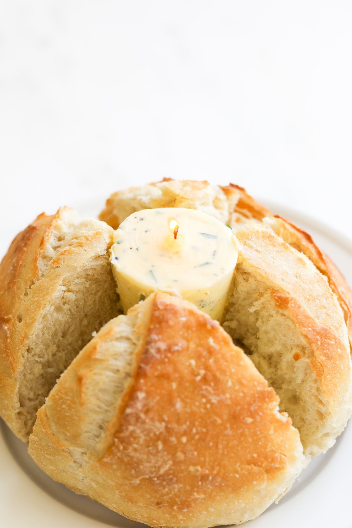 A round loaf of bread is scored into sections, revealing a lit herb butter candle in the center. The dish is placed on a white plate.