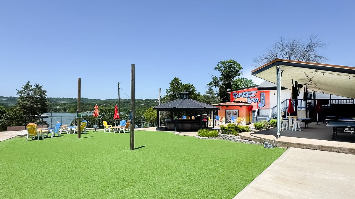 Outdoor bar and seating area with artificial grass, colorful chairs, and umbrellas near a lake—perfect for enjoying views of trees and hills under a clear sky, similar to the inviting vibes found at Table Rock Lake restaurants.