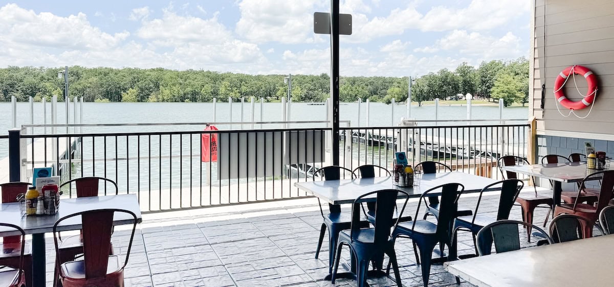 Outdoor dining area with empty tables and chairs overlooking a lake and docks, surrounded by trees and a red life preserver—perfect for those seeking the scenic charm found at Table Rock Lake restaurants.