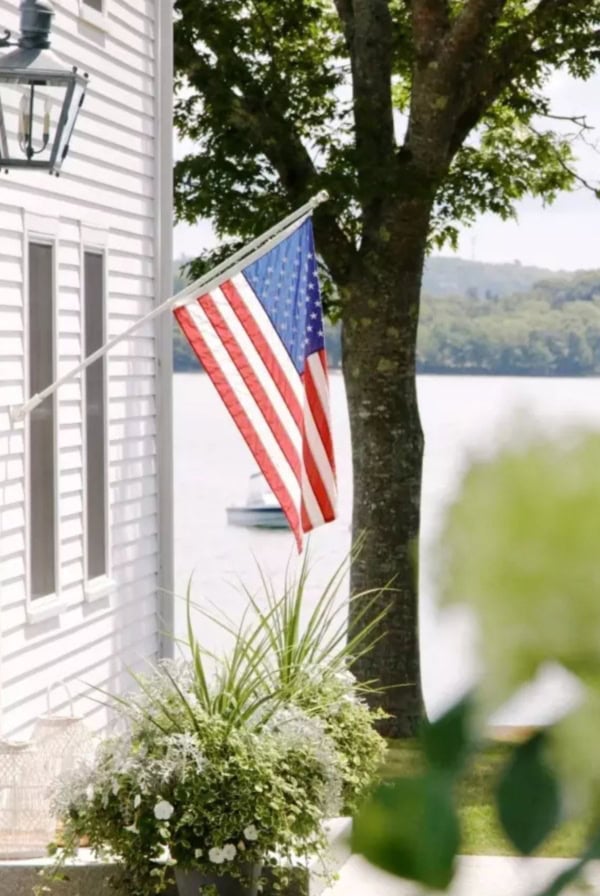 An American flag is mounted on the side of a white house near a lake, with a boat visible on the water and trees in the background.